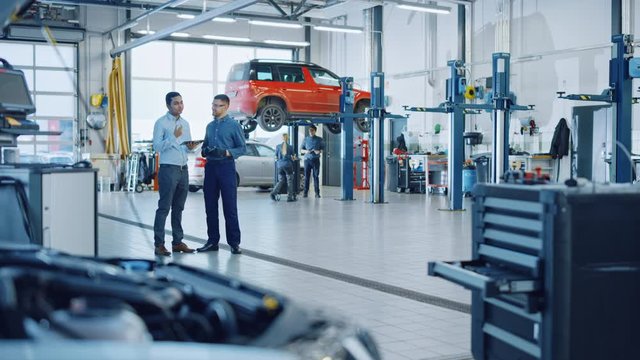 Manager Checks Diagnostics Results on a Tablet Computer and Explains a Vehicle Breakdown to a Handsome Mechanic. Car Service Employees Talk while Standing in a Garage. Modern Clean Workshop.