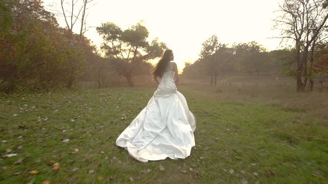 Beautiful brunette bride in long bridal dress running on sunset along fantastic autumn colorful forest. Slow Motion