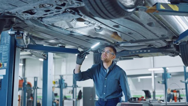 Handsome Professional Car Mechanic is Investigating Rust Under a Vehicle on a Lift in Service. Repairman is Using a LED lamp and Walks Towards. Specialist is Wearing Safety Glasses. Modern Workshop.
