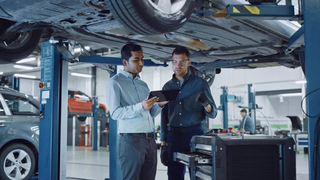 Manager Checks Data on a Tablet Computer and Explains the Breakdown to a Mechanic. Car Service Employees Inspect the Bottom and Skid Plates of the Car. Modern Clean Workshop.