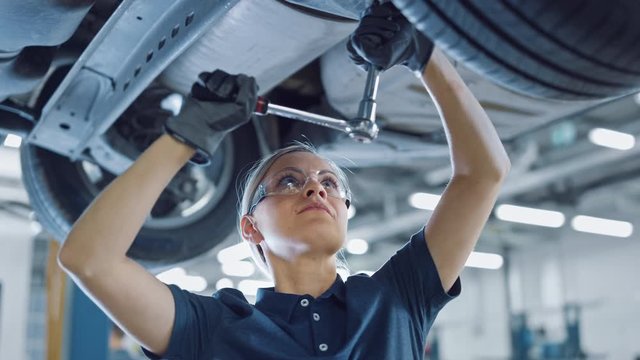 Slow Motion Portrait Footage of a Female Mechanic Working on a Vehicle in a Car Service. Empowering Woman Wearing Gloves and Using a Ratchet Underneath the Car. Modern Clean Workshop. 
