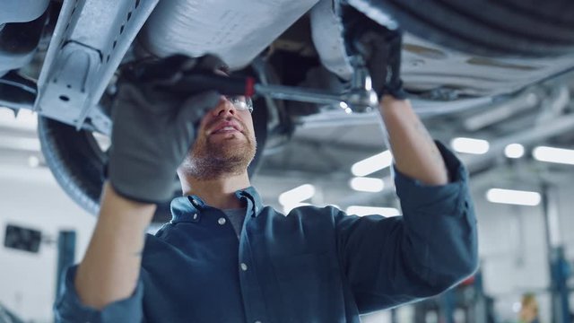 Portrait Footage of a Handsome Mechanic Working on a Vehicle in a Car Service. Professional Repairman is Wearing Gloves and Using a Ratchet Underneath the Car. Modern Clean Workshop. 