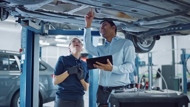 Female Mechanic Talking to a Manager Under a Vehicle in a Car Service. Specialist is Showing Info on a Tablet Computer. Empowering Woman Wearing Gloves and Safety Gloves. Modern Clean Workshop. 