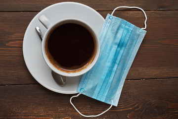 Biohazard concept of living in a new reality of coronavirus epidemy hysteria medical mask and big white mug with a black coffee on wooden table