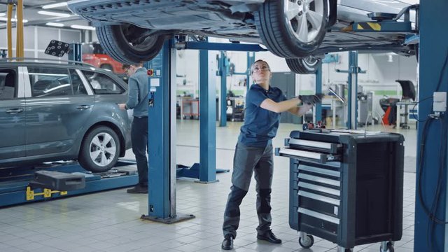 Beautiful Professional Female Car Mechanic is Working Under a Vehicle on a Lift in Service. She is Using a LED lamp and Takes a Ratchet. Specialist is Wearing Safety Glasses. Modern Clean Workshop.