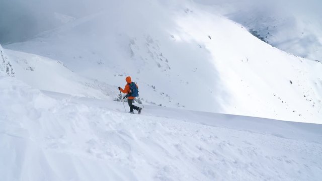 High mountaineer backpacker dressed bright orange softshell jacket using a trekking poles descending the snowy mountain summit. Active people 4K concept footage