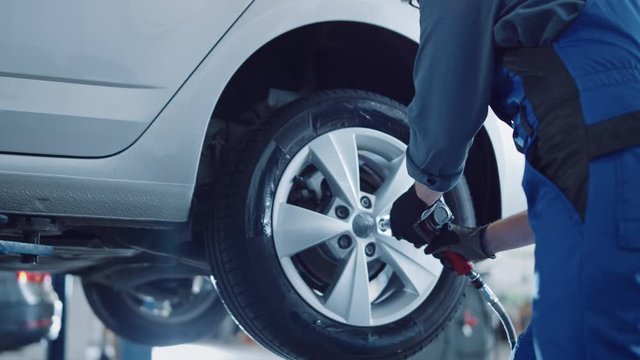 Mechanic in Blue Overalls is Unscrewing Lug Nuts with a Pneumatic Impact Wrench. Repairman Works in a Modern Clean Car Service. Specialists Removes the Wheel in Order to Fix a Component on a Vehicle.