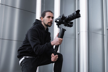 Young Professional videographer holding professional camera on 3-axis gimbal stabilizer. Pro equipment helps to make high quality video without shaking. Cameraman wearing white hoodie making a videos.