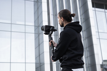 Young Professional videographer holding professional camera on 3-axis gimbal stabilizer. Pro equipment helps to make high quality video without shaking. Cameraman wearing white hoodie making a videos. © Volodymyr_sh