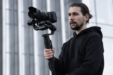 Young Professional videographer holding professional camera on 3-axis gimbal stabilizer. Pro equipment helps to make high quality video without shaking. Cameraman wearing white hoodie making a videos.