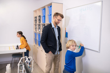 Male teacher standing at whiteboard, boy writing, girl playing with building kit