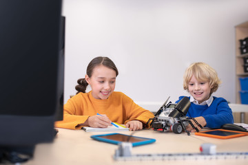 Pupils sitting at desk, taking notes, playing with building kit, smiling