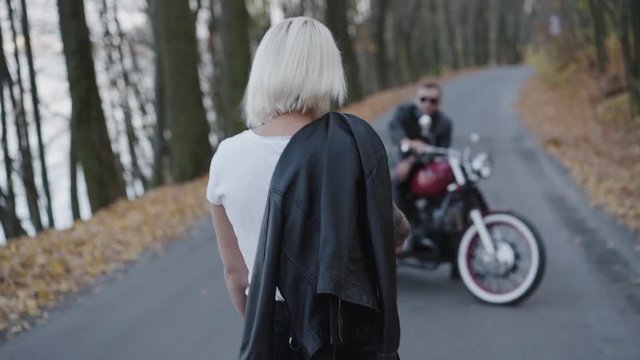 Back look of blonde comes to her smiling biker sitting at motorcycle in park