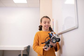 Girl standing at whiteboard, holding buildable car, looking at it