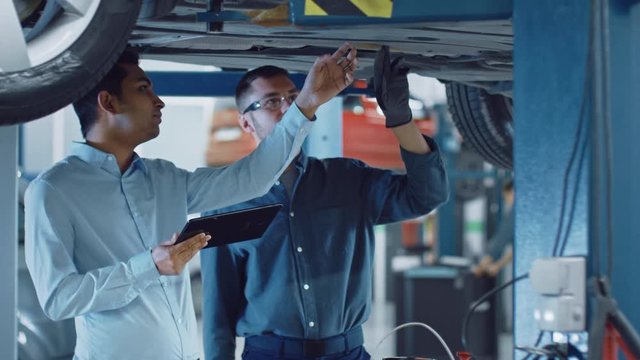 Manager Checks Data on a Tablet Computer and Explains the Breakdown to a Mechanic. Car Service Employees Inspect the Bottom and Skid Plates of the Car. Modern Clean Workshop.