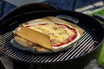 A portrait of a pizza margarita lying on a pizza stone and a piece of baking paper on a charcoal barbecue grill. The delicious pizza has cheese and tomato sauce on it.
