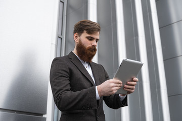 Modern Young bearded Business man working with a digital tablet. Young hipster businessman holding tablet in hands outdoor. Working online with a tablet while standing outside on an office building.