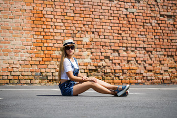 Fototapeta na wymiar Girl in denim overalls, a white T-shirt and a light hat on a background of a brick building under the bright sun