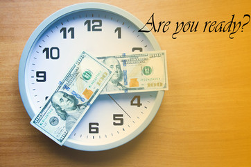 Analog clock and dollars on a wooden background. Banknotes are lying on the clock in the form of arrows. 100 dollars indicate hours and minutes. View from above. On the clock  time 20:15, 08:15