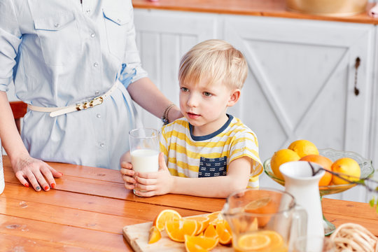 The Boy Drinks Milk From A Glass. Mother And Son Are Smiling While Having A Breakfast In Kitchen. Mom Is Pouring Milk Into Glass