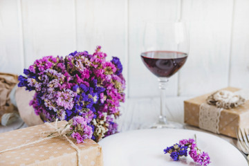 Table setting. Plate on wooden background with gift box, cutlery, a glass of red wine and flowers.