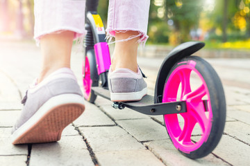 Bottom part of young adult female person wearing pink casual jeans and sneakers riding push kick scooter at city street on summer day outdoor. Teenager girl having fun enjoy sport activities outside