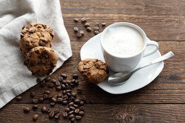 A white cup of cappuccino, coffee with froth on a saucer, coffee beans and cookies biscuits nearby. Cookies lie on a linen linen napkin on a rustic wooden table. Breakfast. Dessert. morning in cafe
