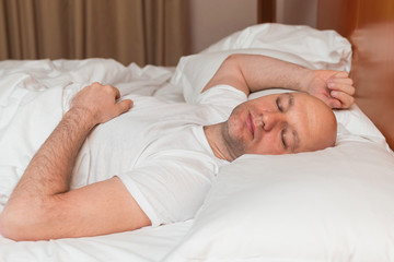 Bald man in white t shirt sleeps in a bed,