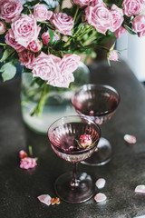 Valentines day holiday greeting card. Champagne in glasses and tender pink roses bouquet in vase at background, selective focus. Romantic couple celebration