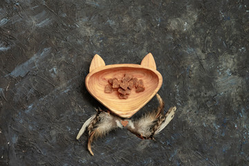 Wooden dish with a treat for petsshaped in head cat and feathers of cat toy on dark background. View from above. Nutrition and pet care concept