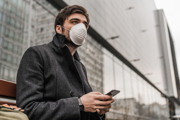 Handsome man sitting on the bench wears mask to prevent virus and for protection while using mobile...