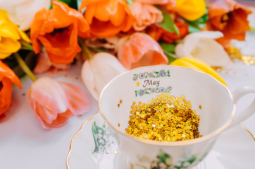 many Golden spangles of stars in white porcelain mug with text may month on a smooth light white background. Bouquet of multi-colored tulips on the background.