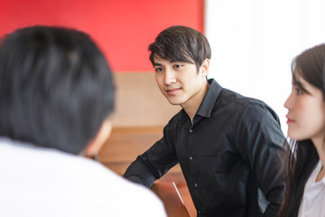 asian businessman sitting around a table having a meeting, talking to each other on information, data and statistics, wearing a black long sleeves shirt and sitting next to an asian businesswoman.