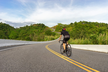 back view of a cyclist on top of a mountains winding road, riding a black bicycle down the wearing...