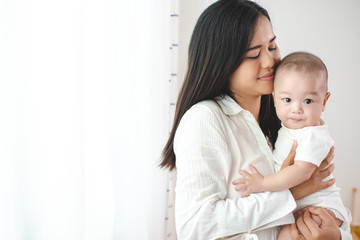 Asian mother holding his child happily. In the bedroom with white curtains. Motherhood, family relationship, new born baby, love of mother to her son or daughter concept.