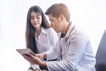 asian male doctor wearing a white scrub lab coat with a stethoscope around his neck smiling and holding a tablet showing health information to a young beautiful asian female patient smartly dressed.