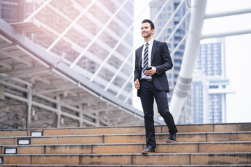 Caucasian handsome businessman confidently walking down the stairways while holding a smartphone, wearing black suit and tie with city skylines and urban architectural structure in the background