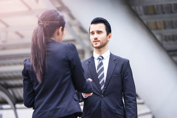 caucasian businessman smiling and standing casually shaking hands with a beautiful asian businesswoman in greetings, outdoor within the city district with urban architectural structure in background