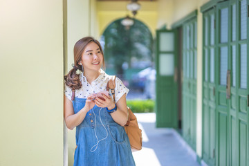 asian women tourist listening to music by playing on the phone with earphones in, smiling leaning against a pillar in the hallway with suns shining and wearing a denim maternity and a brown bag pack