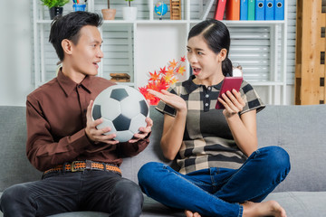 Good looking young asian couple looking surprised while sitting on sofa and watching soccer game on tv at home.