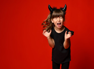 Little kid with devil horns, in black blouse and leggings screaming with closed eyes, posing...