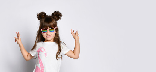 A small cheerful girl, in dark glasses in a White Studio. Stands with his hands raised and shows the peace gesture.