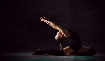 Young girl gymnast in black sport body and special footwear making gymnastic pose over dark background