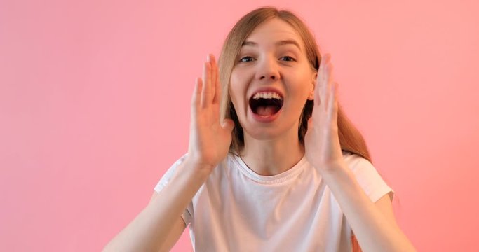 A young shocked woman in a white t-shirt , holding her hand near her mouth and telling a Secret, on a pink background, a girl screams