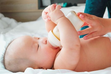 Portrait of a newborn Asian baby on the bed drinks milk from a bottle, Charming black-eyed baby 4 month old lies in bed ,A child resting on a bed