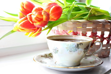 White porcelain pattern cup of hot black tea on dish on white desk on blurred Bouquet of flowers of red yellow blooming tulips with green leaf in wicker basket. Selective focus