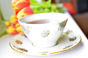 White porcelain pattern cup of hot black tea on dish on white desk in room with blurred Bouquet of flowers of red yellow blooming tulips with green leaf on backdrop. Selective focus, tilt frame