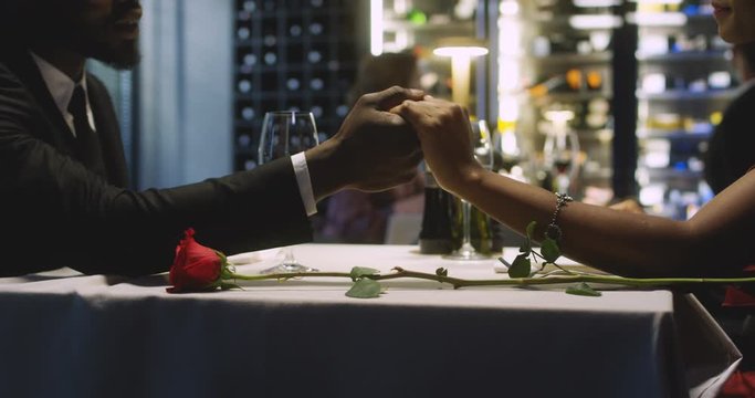 Close up of African American male and female hands touching and holding while lying on table next to red rose at dinner in restaurant. Romantic supper with white wine. Couple in love at date concept.