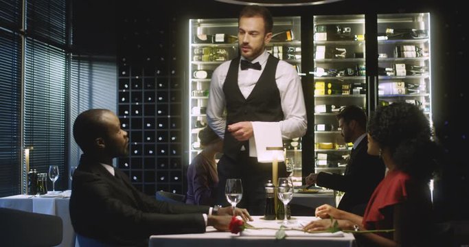 Caucasian young male waiter coming to table of African American couple clients and talking about order. Happy man and woman ordering dinner in restaurant. Good service concept.