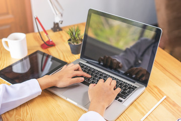 close up of a doctor typing on the keyboard of the laptop computer with both hands, researching work or diagnosis patients health, working in an office on the wooden desk with tablet and stethoscope